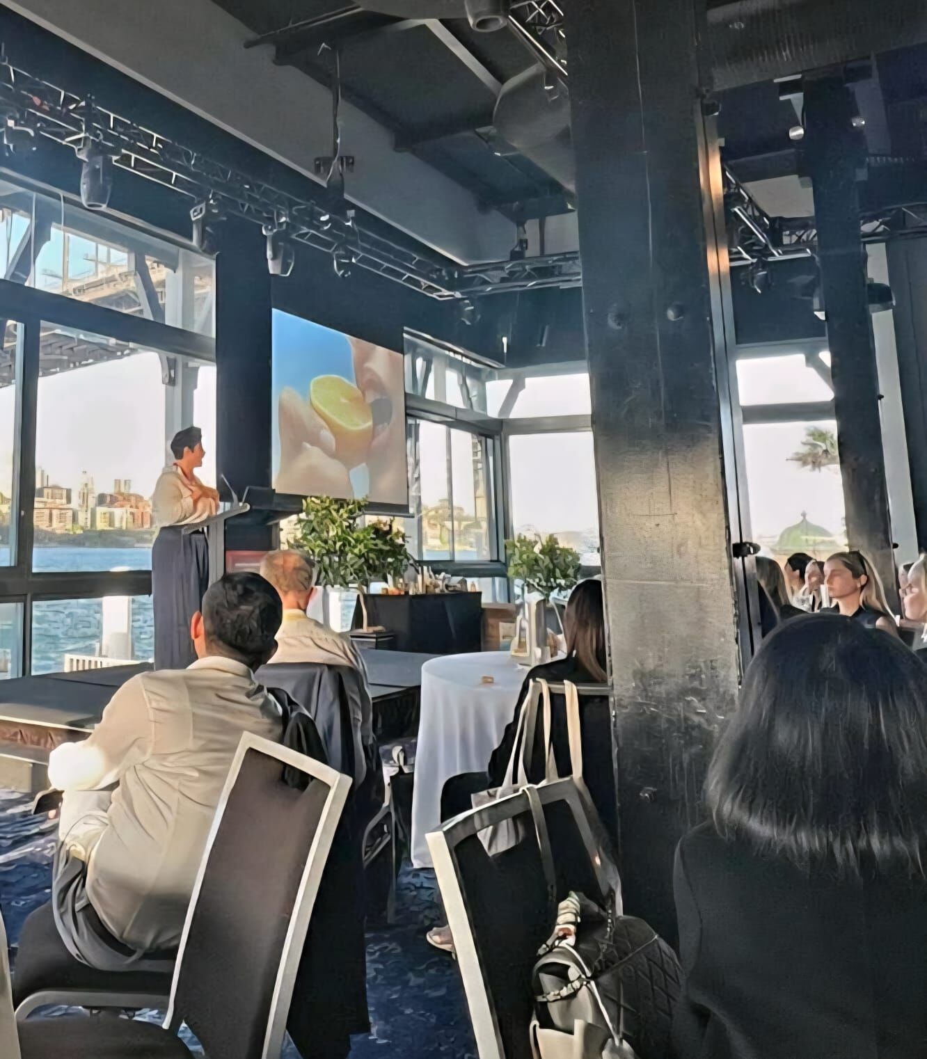 Amanda Anderson presenting at a waterfront retail industry event — audience seated, large screen behind her